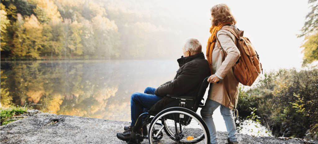 woman pushes elderly man in wheelchair next to a body of water outside