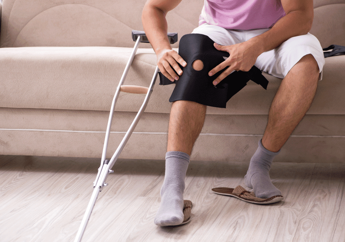 Man sitting on a couch adjusting a black knee brace with crutches beside him