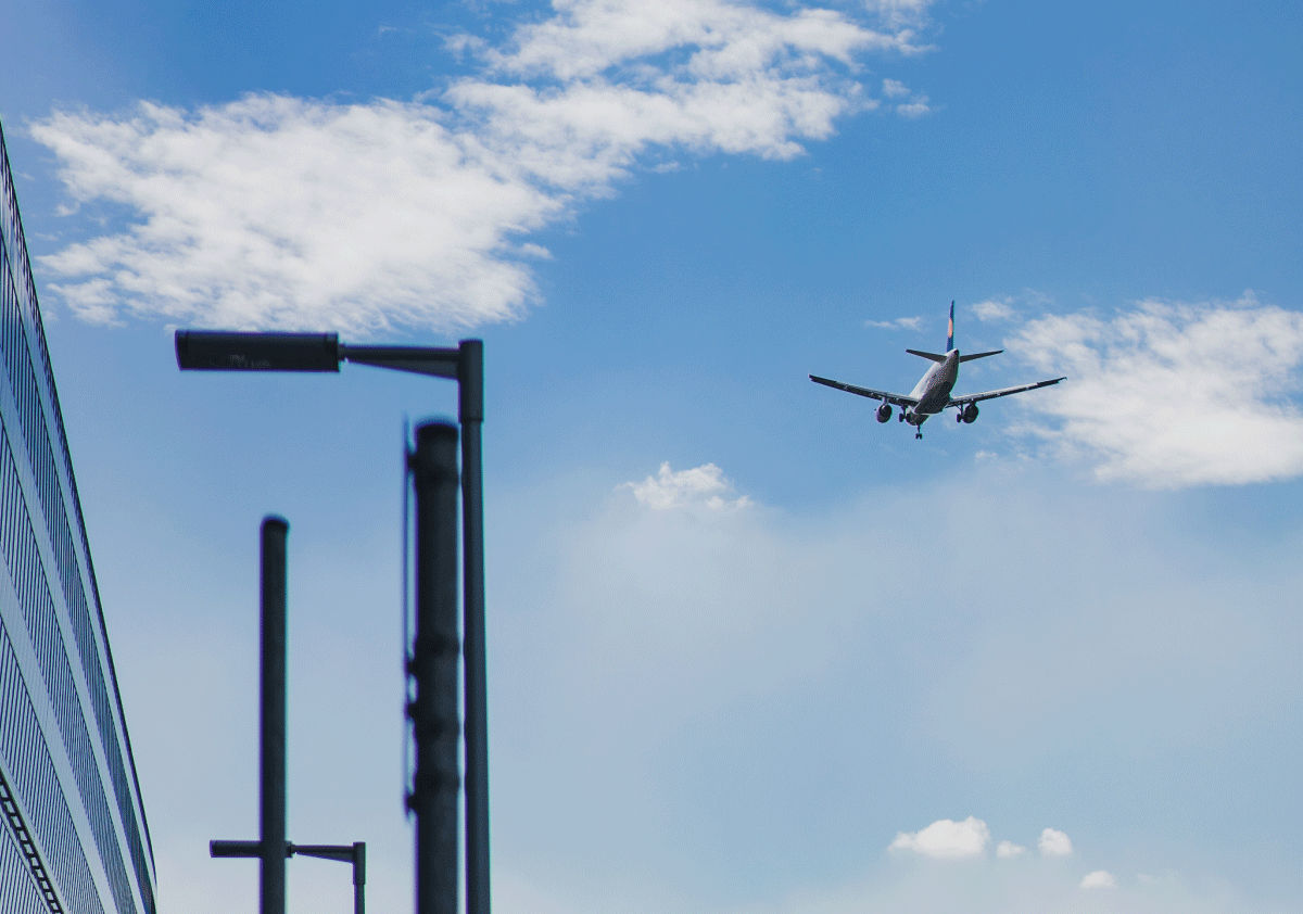 Airplane flying in a clear blue sky near buildings, symbolizing air travel.