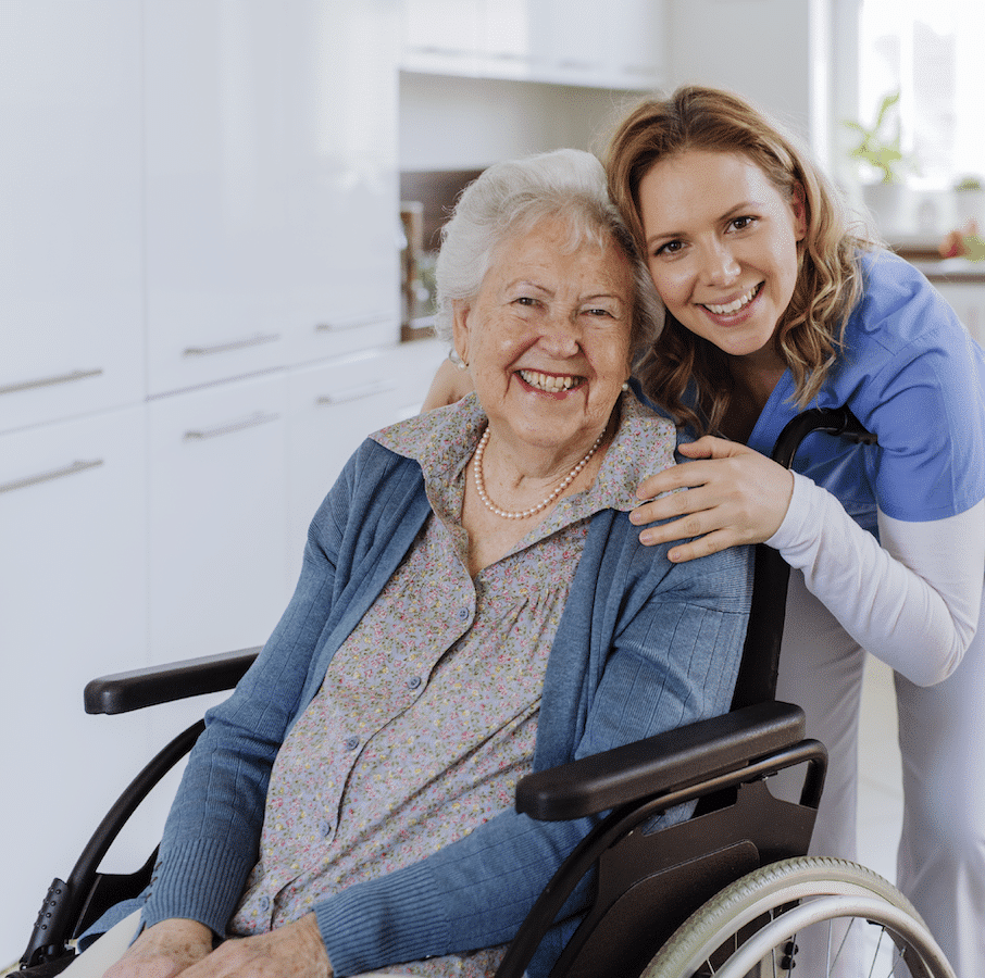 An elderly woman in a wheelchair and a younger woman in scrubs smiling at the camera