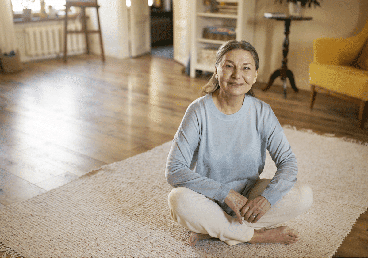 An elderly woman smiling and sitting cross-legged on her cozy rug in comfort thanks to her incontinence products from Alpine Home Medical